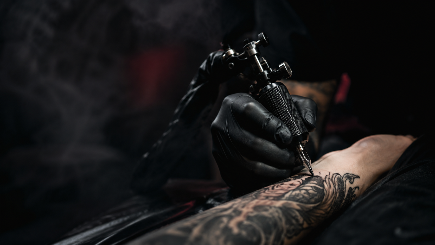 Black gloved hand holding a tattoo machine over a forearm in a dark cinematic craft scene